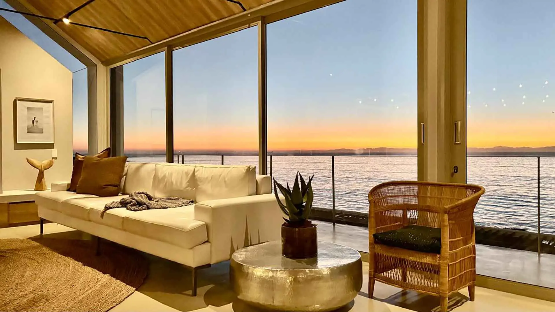 Lounge at sunset with a white sofa and rattan chair, the bay turning orange outside
