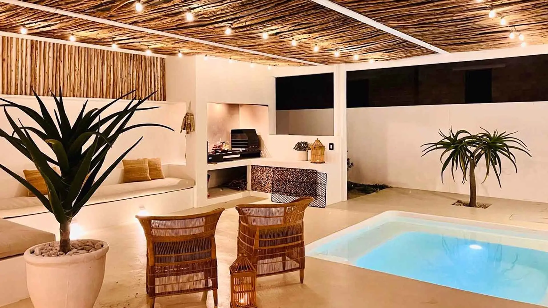 Courtyard at night: the plunge pool lit blue under a pergola strung with festoon lights, the braai alight
