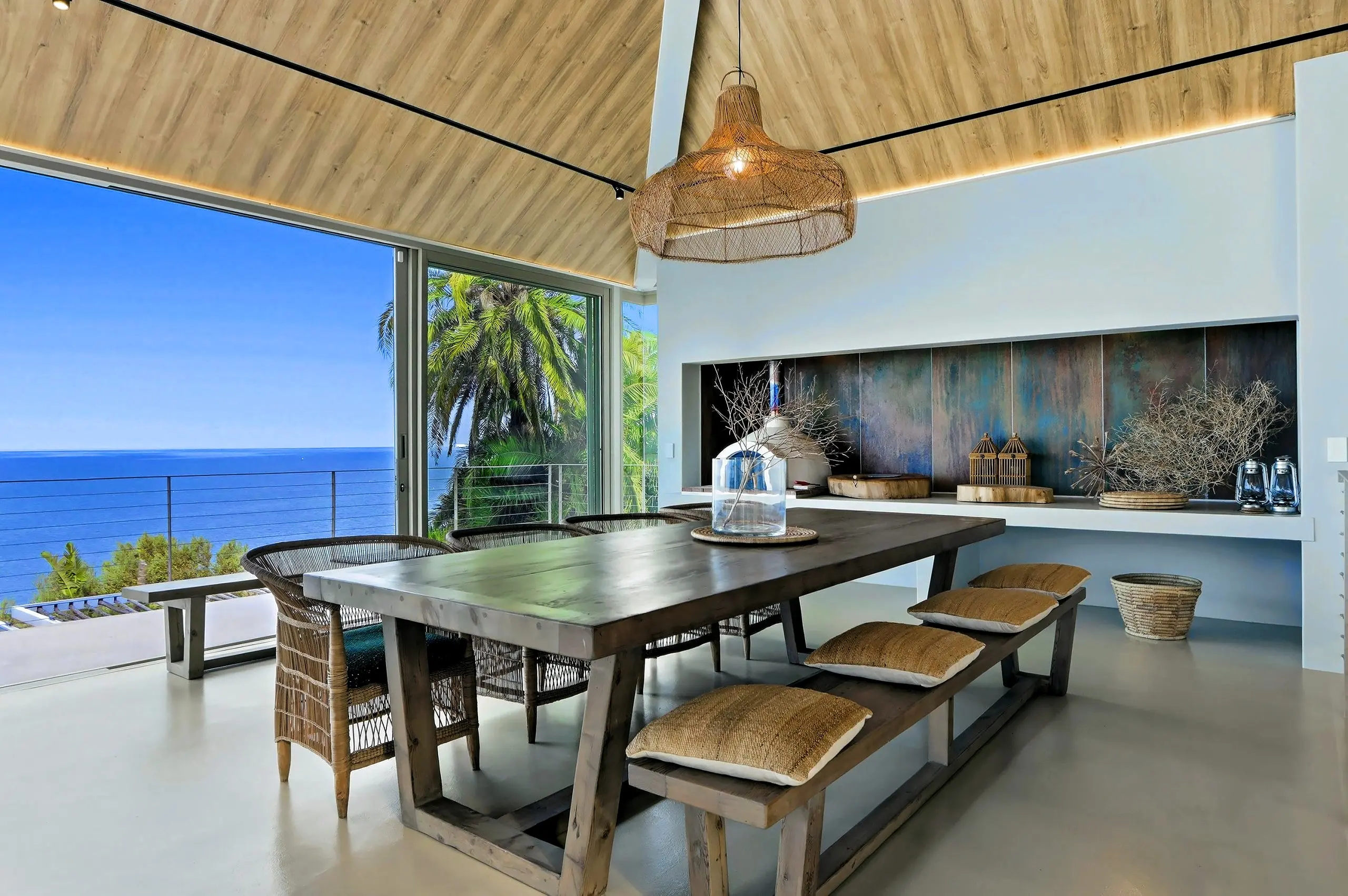 Harvest dining table beside sliding glass to the ocean, with a woven pendant and a copper feature wall