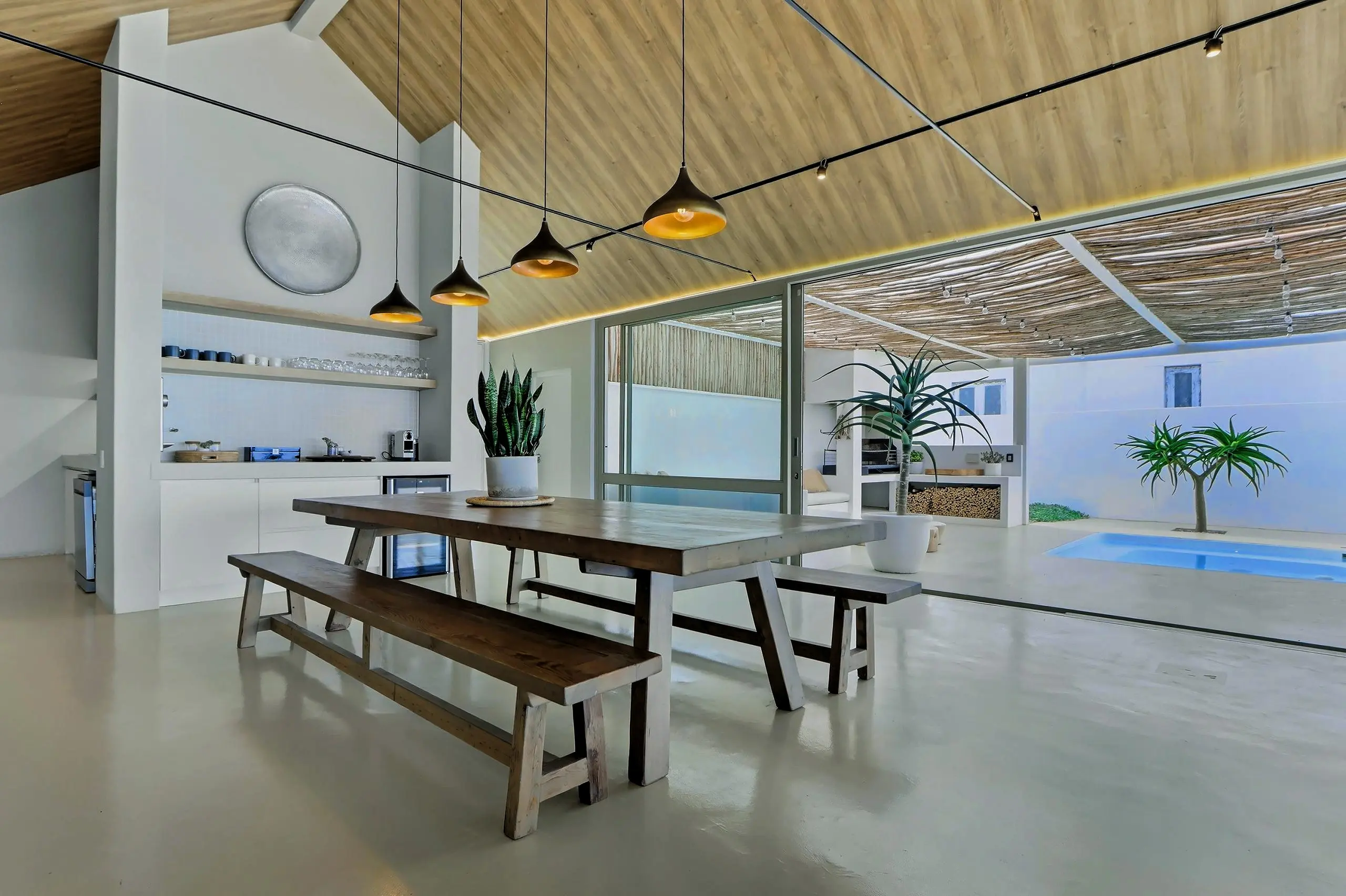 Second dining table with benches under brass pendants, the courtyard pool beyond the glass