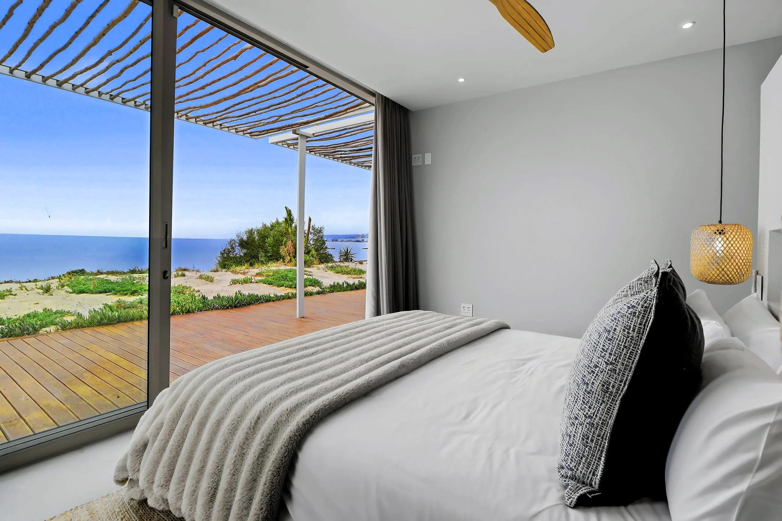 Bedroom with a grey throw, bamboo pendant and glass doors to a timber deck and the ocean