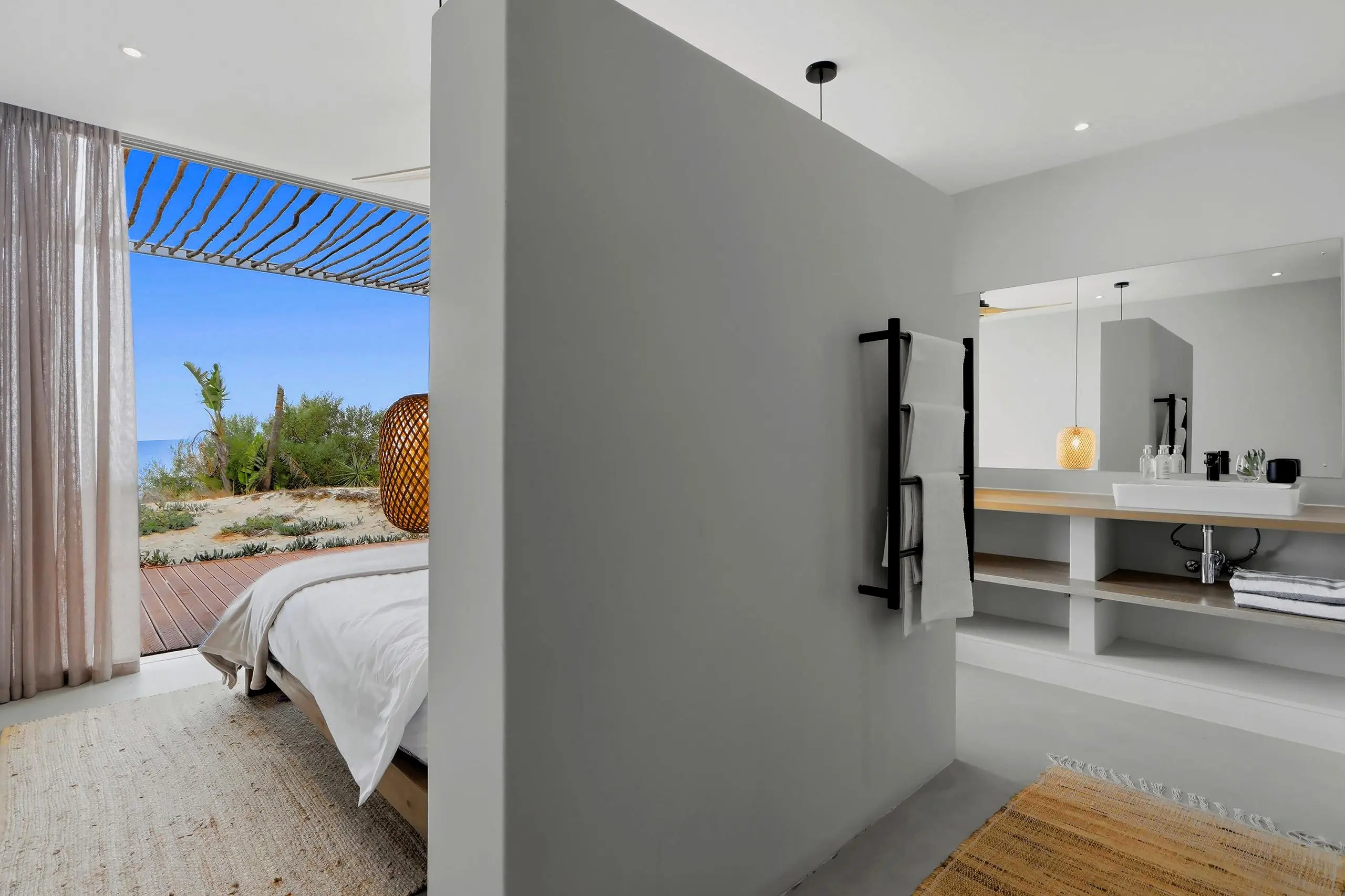 Bedroom with a bed facing the deck and pergola, vanity and heated towel rail behind a half wall