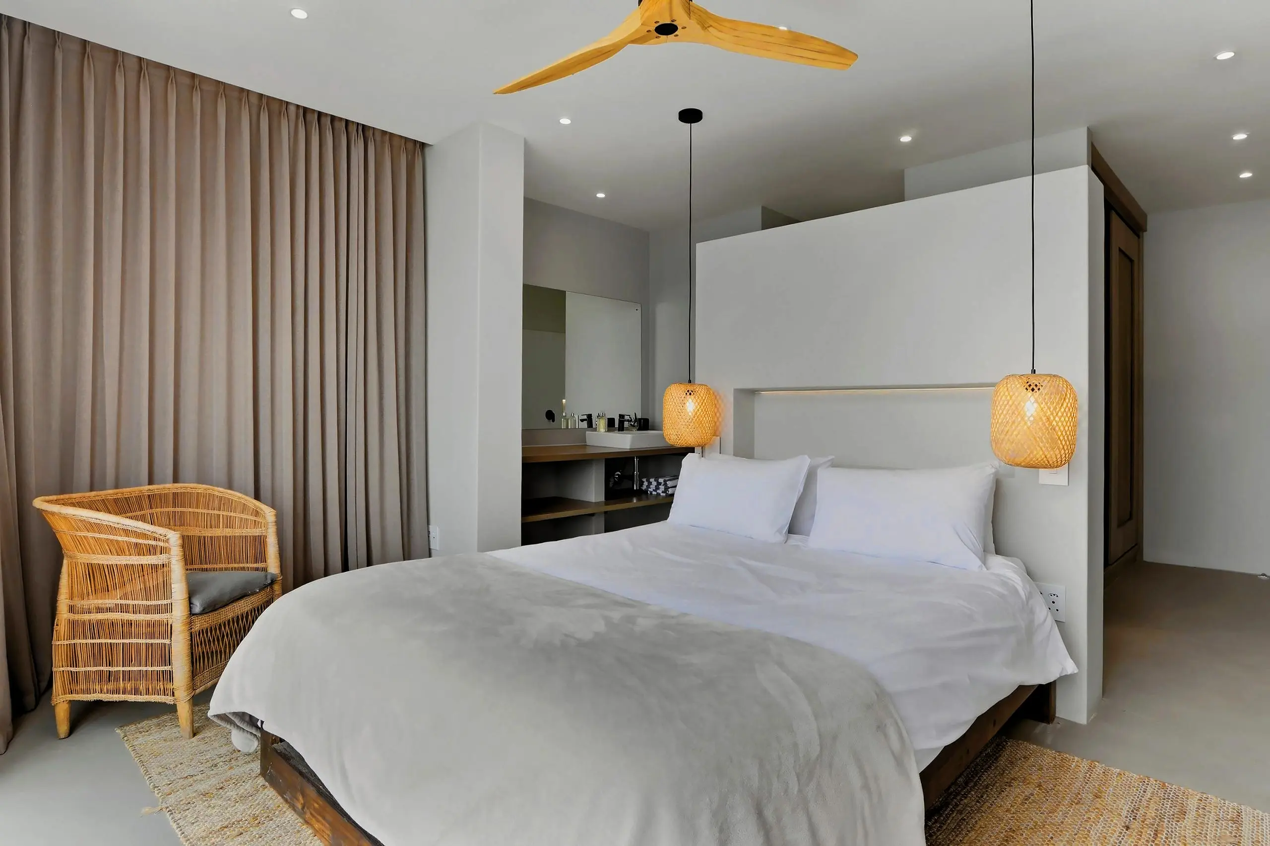 Bedroom with linen curtains, rattan chair, bamboo pendants and a vanity behind the headboard wall
