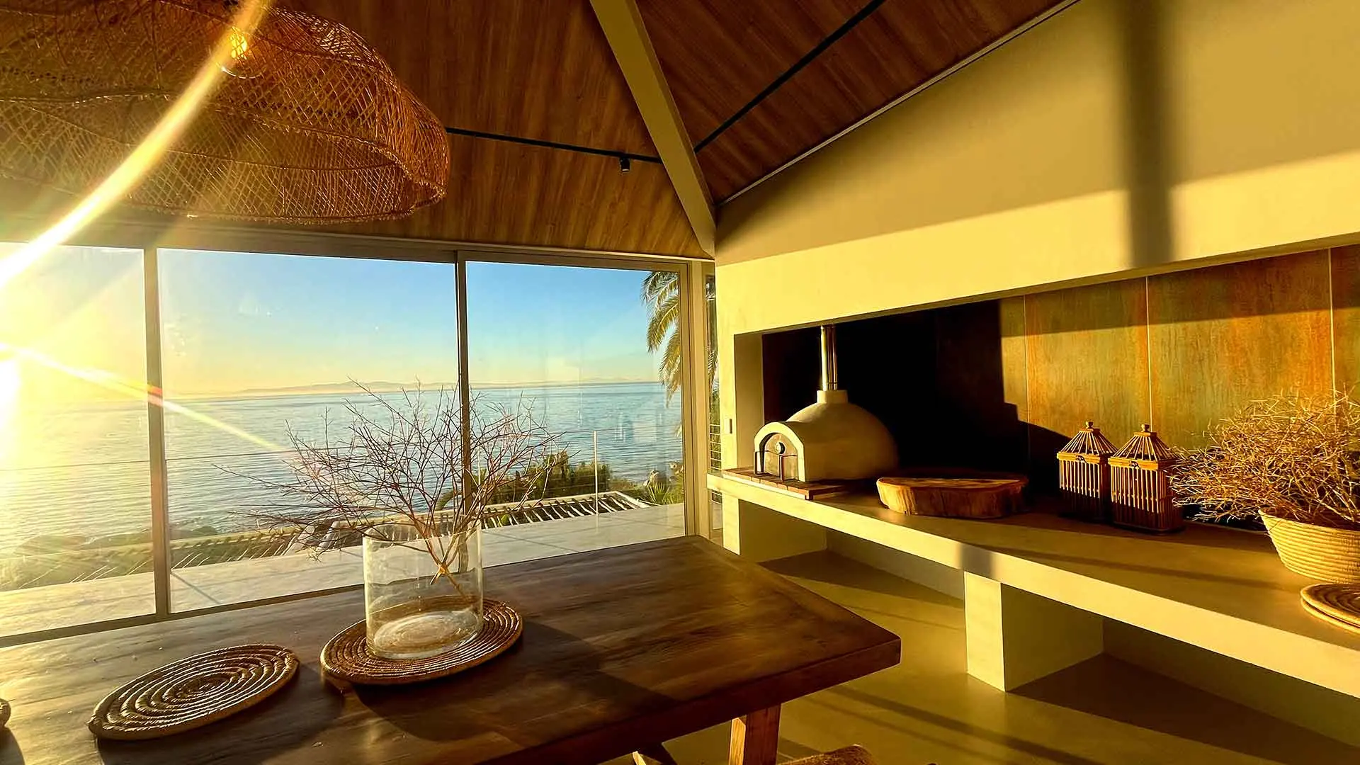 Dining table at sunrise with the pizza oven and copper wall, the sea pale beyond the glass