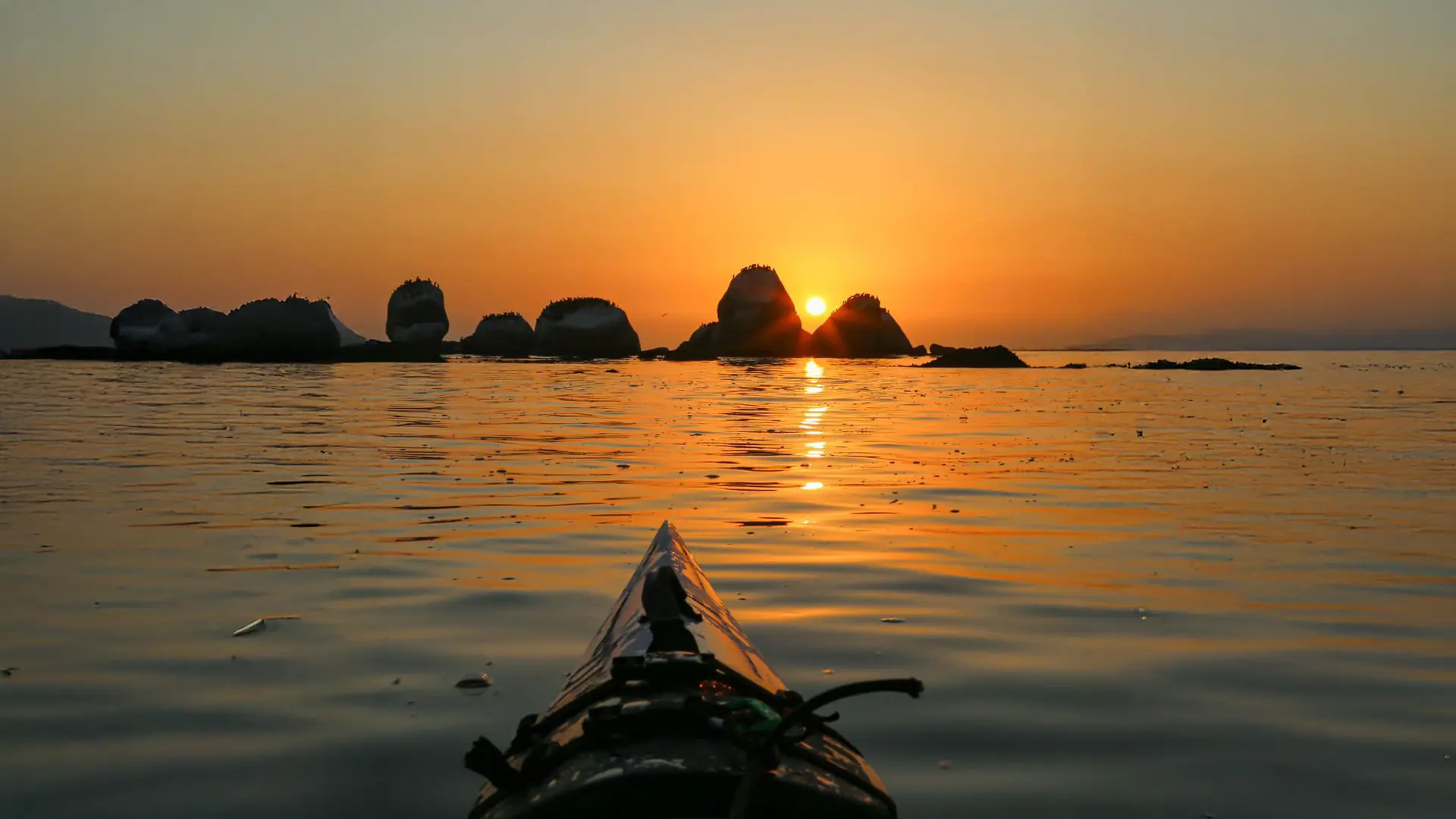 Kayak bow on glassy water at sunrise, the sun rising between rocks in the bay