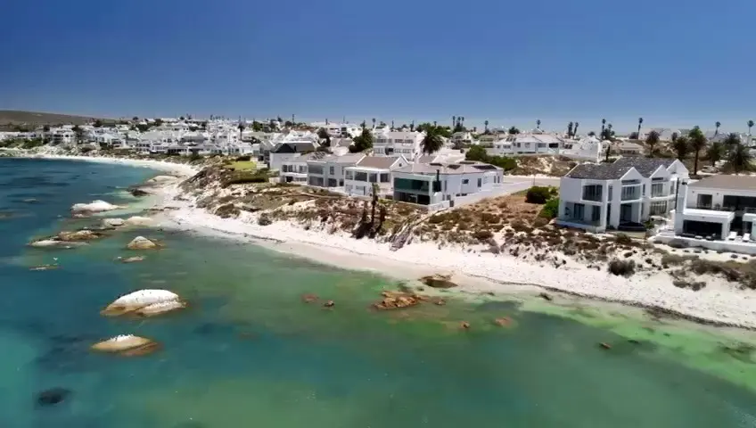 Still from the Villa Selah film: the white villa on the shore of the bay from the air, beach and boulders below
