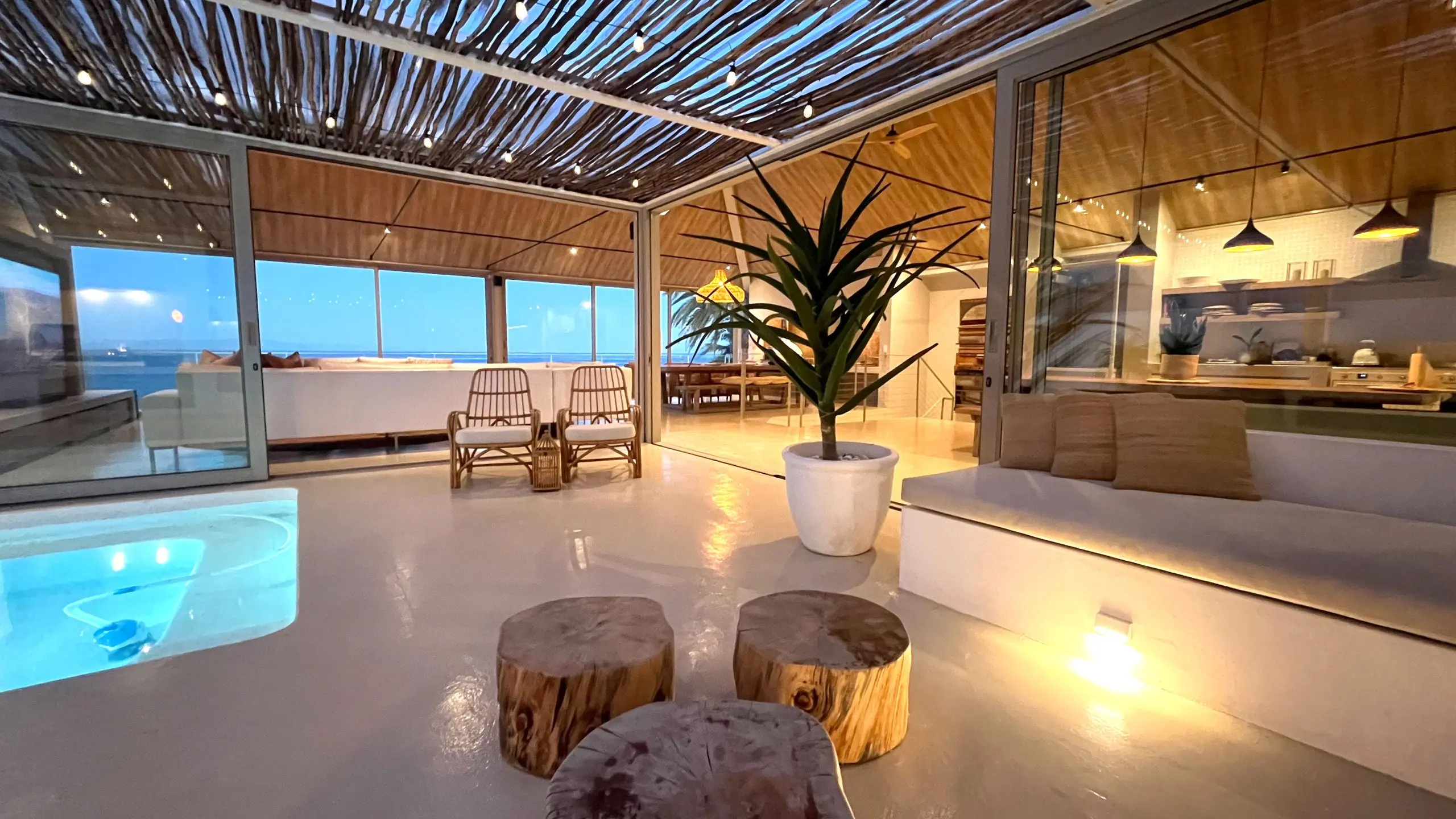 Pool terrace at dusk under a latte-wood pergola strung with lights, the lounge lit behind